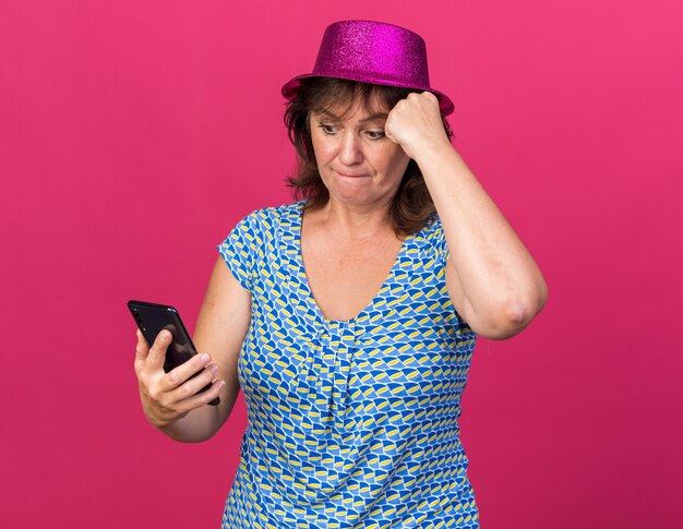 Mujer de mediana edad con gorro de fiesta mirando la pantalla de su teléfono inteligente confundida celebrando la fiesta de cumpleaños de pie sobre la pared rosa