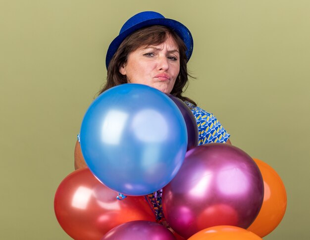 Mujer de mediana edad con gorro de fiesta con un montón de globos de colores con el ceño fruncido