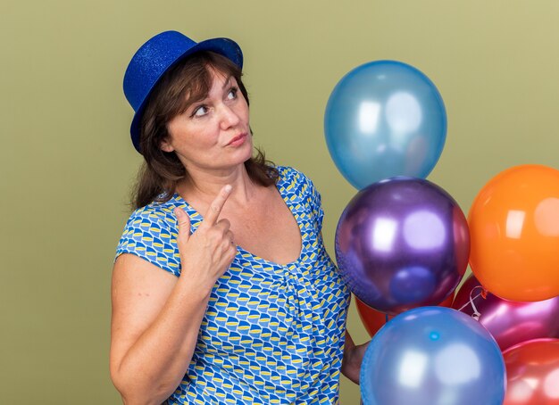 Mujer de mediana edad con gorro de fiesta con un montón de globos de colores mirando desconcertado celebrando la fiesta de cumpleaños de pie sobre la pared verde