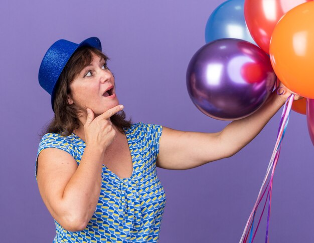 Mujer de mediana edad con gorro de fiesta con un montón de globos de colores mirándolos asombrados y sorprendidos celebrando la fiesta de cumpleaños de pie sobre la pared púrpura