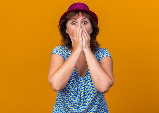 Mujer de mediana edad con gorro de fiesta sorprendido cubriendo la boca con las manos celebrando la fiesta de cumpleaños de pie sobre la pared naranja