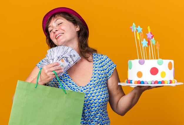 Mujer de mediana edad con gorro de fiesta sosteniendo una bolsa de papel con regalos sosteniendo pastel de cumpleaños y dinero en efectivo feliz y complacido sonriendo alegremente