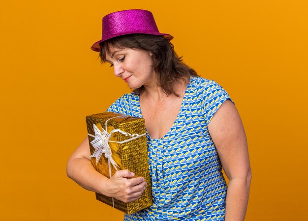Mujer de mediana edad con gorro de fiesta sosteniendo un presente mirando hacia abajo con una sonrisa tímida en la cara