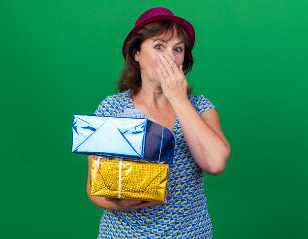 Mujer de mediana edad con gorro de fiesta sosteniendo regalos de cumpleaños conmocionado cubriendo la boca con la mano