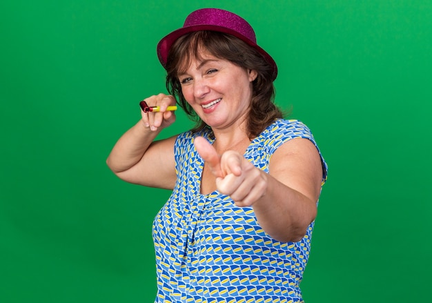 Mujer de mediana edad con gorro de fiesta sosteniendo un silbato apuntando con el dedo índice sonriendo alegremente celebrando la fiesta de cumpleaños de pie sobre la pared verde