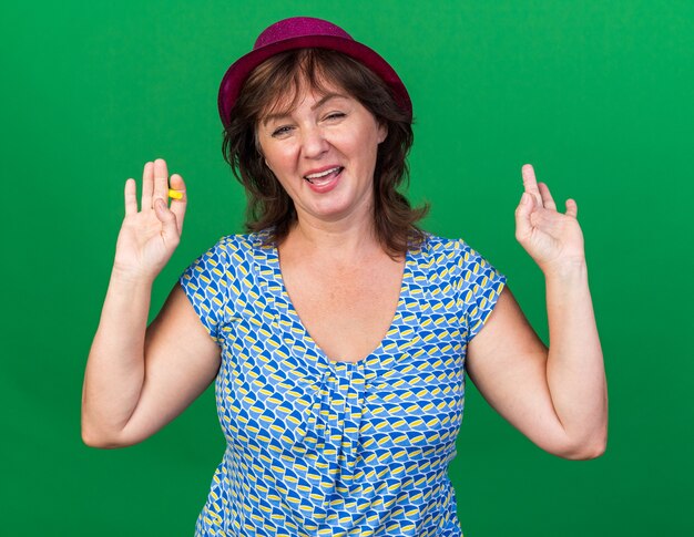 Mujer de mediana edad con gorro de fiesta sosteniendo un silbato feliz y alegre con los brazos levantados celebrando la fiesta de cumpleaños de pie sobre la pared verde