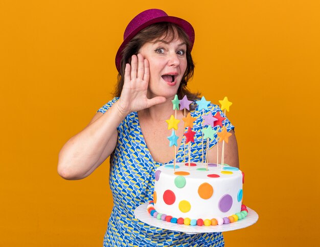 Mujer de mediana edad con gorro de fiesta sosteniendo la torta de cumpleaños con la mano sobre la oreja tratando de escuchar chismes