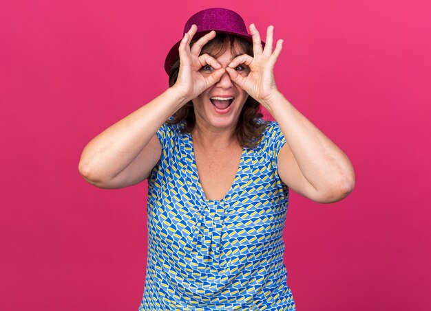 Mujer de mediana edad con gorro de fiesta a través de los dedos haciendo gesto binocular sonriendo alegremente