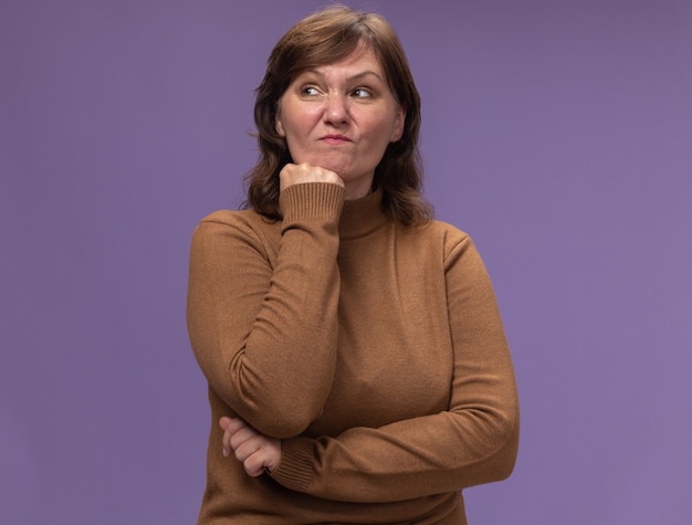 Mujer de mediana edad insatisfecha en cuello alto marrón mirando a un lado con la mano en la barbilla pensando de pie sobre la pared púrpura