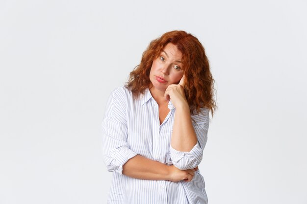 Mujer de mediana edad molesta y molesta, cansada con cabello rojo que parece agotada y harta, apoyándose en la mano y mirando a la cámara escéptica, escuchando conversaciones sin sentido, fondo blanco.