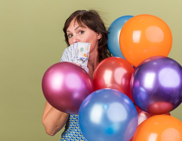 Mujer de mediana edad montón de globos de colores con efectivo feliz y sorprendido celebrando la fiesta de cumpleaños de pie sobre la pared verde