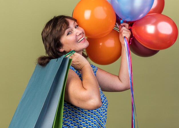 Mujer de mediana edad con un montón de globos de colores sosteniendo bolsas de papel con regalos feliz y emocionado celebrando la fiesta de cumpleaños de pie sobre la pared verde