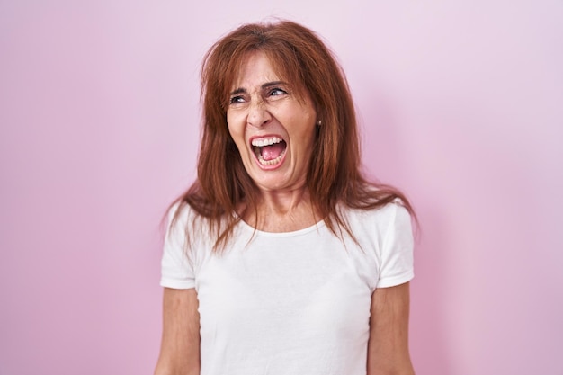 Mujer de mediana edad parada sobre un fondo rosado enojada y loca gritando frustrada y furiosa, gritando con ira. rabia y concepto agresivo.