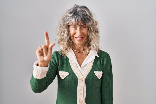 Mujer de mediana edad de pie sobre fondo blanco que aparece y señala con el dedo número uno mientras sonríe confiada y feliz.