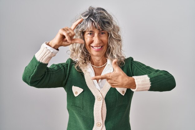 Mujer de mediana edad de pie sobre fondo blanco sonriendo haciendo marco con manos y dedos con cara feliz. concepto de creatividad y fotografía.