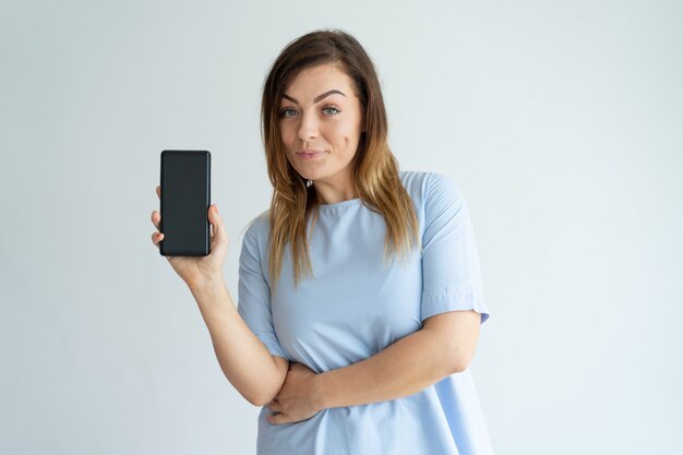 Mujer de mediana edad positiva que muestra la pantalla del teléfono inteligente y mirando a cámara.