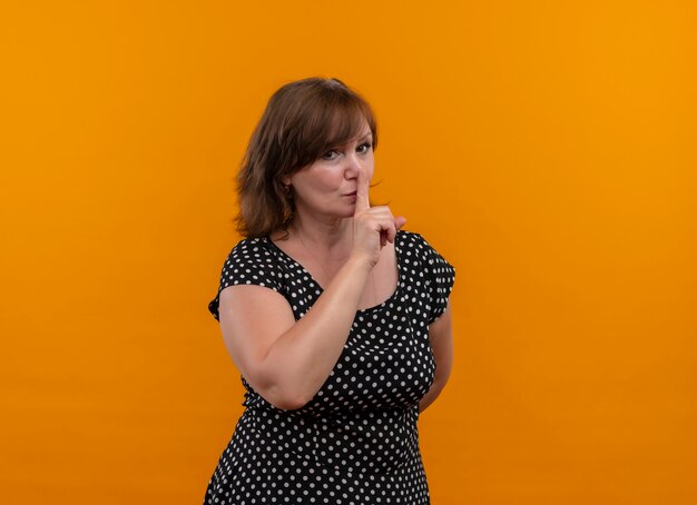 Mujer de mediana edad que mira seriamente que gesticula el silencio en la pared anaranjada aislada con el espacio de la copia