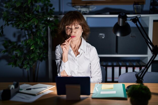 Mujer de mediana edad que trabaja en la oficina por la noche con la boca y los labios cerrados como zip con los dedos. secreto y silencioso, tabú hablando