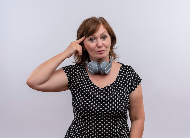 Mujer de mediana edad seriamente mirando poniendo su dedo en la sien con auriculares alrededor de su cuello en la pared blanca aislada