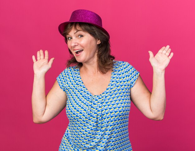 Mujer de mediana edad con sombrero de fiesta feliz y emocionado levantando los brazos