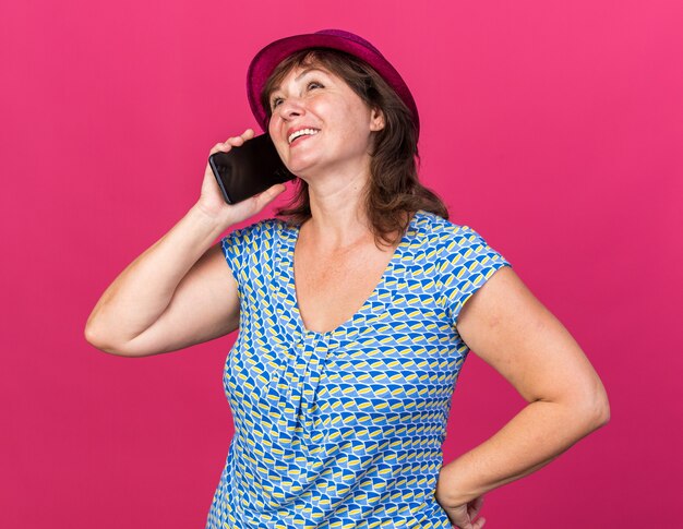 Mujer de mediana edad con sombrero de fiesta feliz y positivo sonriendo alegremente mientras habla por teléfono móvil celebrando la fiesta de cumpleaños de pie sobre la pared rosa