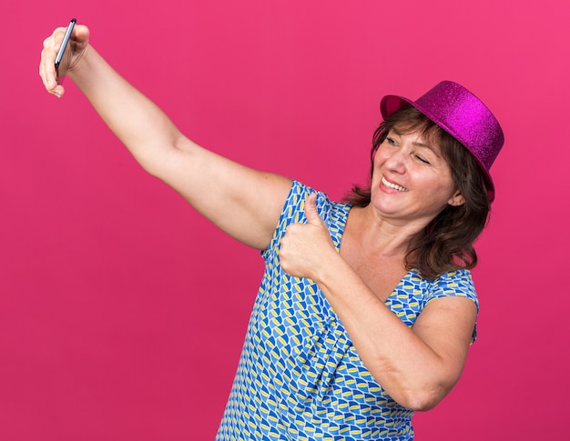 Foto gratuita mujer de mediana edad con sombrero de fiesta haciendo selfie con smartphone sonriendo feliz y alegre mostrando los pulgares para arriba