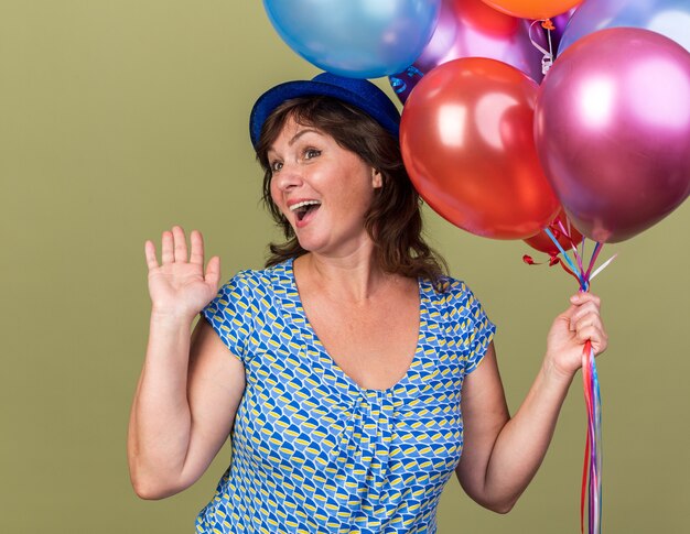 Mujer de mediana edad con sombrero de fiesta con un montón de globos de colores feliz y emocionado sonriendo alegremente con el brazo levantado celebrando la fiesta de cumpleaños de pie sobre la pared verde