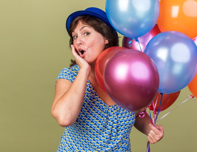 Mujer de mediana edad con sombrero de fiesta con un montón de globos de colores sorprendidos y felices celebrando la fiesta de cumpleaños de pie sobre la pared verde