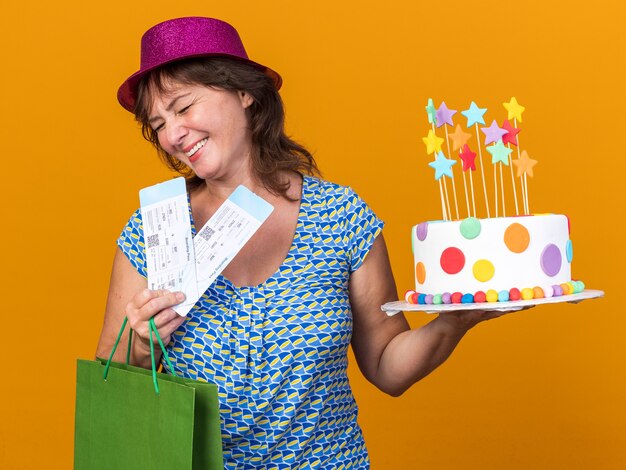 Mujer de mediana edad con sombrero de fiesta sosteniendo una bolsa de papel con regalos sosteniendo pastel de cumpleaños y boletos de avión feliz y complacido sonriendo alegremente celebrando la fiesta de cumpleaños de pie sobre la pared naranja