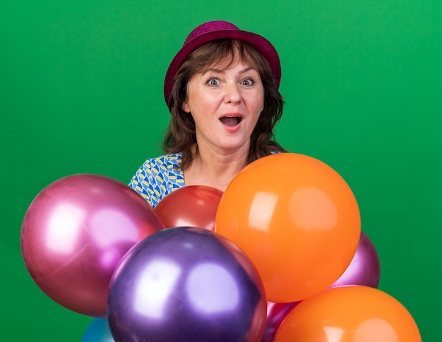 Mujer de mediana edad con sombrero de fiesta sosteniendo globos de colores feliz y emocionado sonriendo alegremente celebrando la fiesta de cumpleaños de pie sobre la pared verde