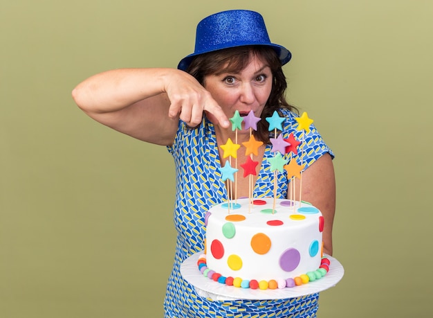 Mujer de mediana edad con sombrero de fiesta sosteniendo pastel de cumpleaños apuntando con el dedo índice feliz y sorprendido celebrando la fiesta de cumpleaños de pie sobre la pared verde