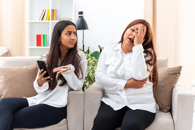 Mujer de mediana edad y su hija joven en camisas blancas y pantalones negros sentados en la hija de sillas con teléfono inteligente peleando con su madre en la sala de luz