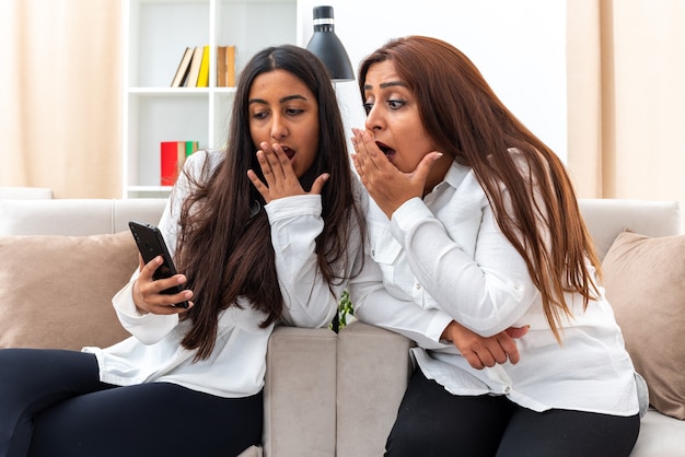 Mujer de mediana edad y su hija joven con camisas blancas y pantalones negros sentados en las sillas hija y su madre con teléfono inteligente mirando asombrados y sorprendidos en la sala de luz