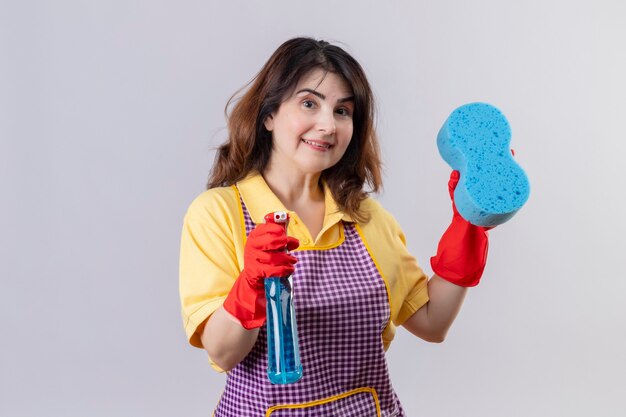 Mujer de mediana edad vistiendo delantal y guantes de goma con esponja y spray de limpieza