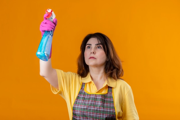 Mujer de mediana edad vistiendo delantal y guantes de goma limpieza con spray de limpieza con rostro serio de pie sobre la pared naranja