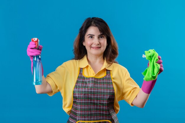 Mujer de mediana edad vistiendo delantal y guantes de goma sosteniendo alfombra y spary de limpieza