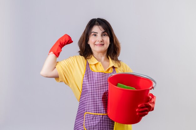 Mujer de mediana edad vistiendo delantal y guantes de goma sosteniendo un balde con herramientas de limpieza mirando a la cámara sonriendo alegremente positivo y feliz levantando el puño regocijándose de su éxito de pie sobre blanco bac