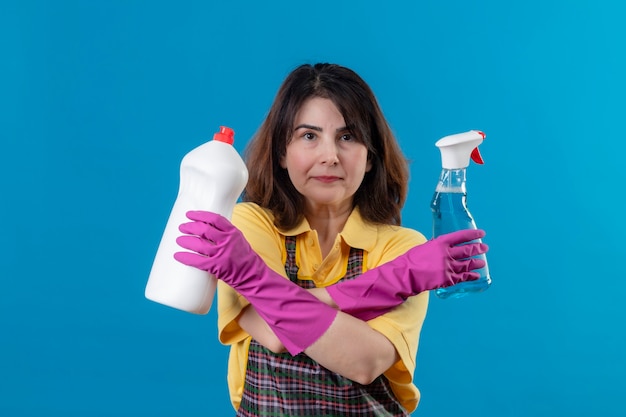 Mujer de mediana edad vistiendo delantal y guantes de goma sosteniendo suministros de limpieza de pie con los brazos cruzados.