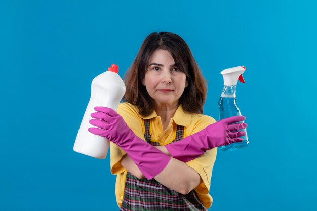 Mujer de mediana edad vistiendo delantal y guantes de goma sosteniendo suministros de limpieza de pie con los brazos cruzados.