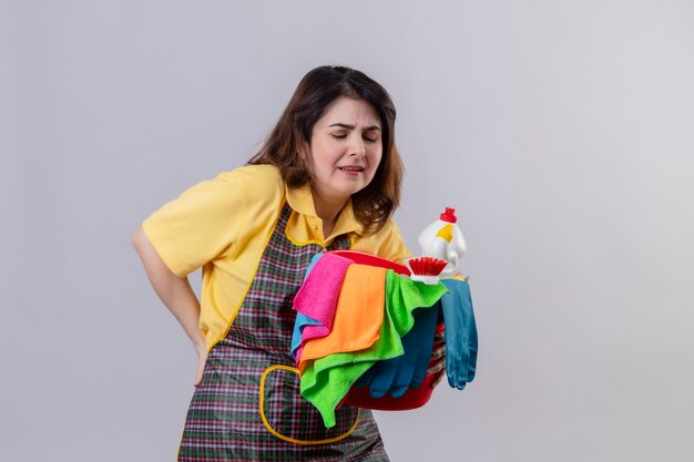 Mujer de mediana edad vistiendo delantal sosteniendo el balde con herramientas de limpieza mirando mal tocando su espalda con dolor de pie sobre la pared blanca