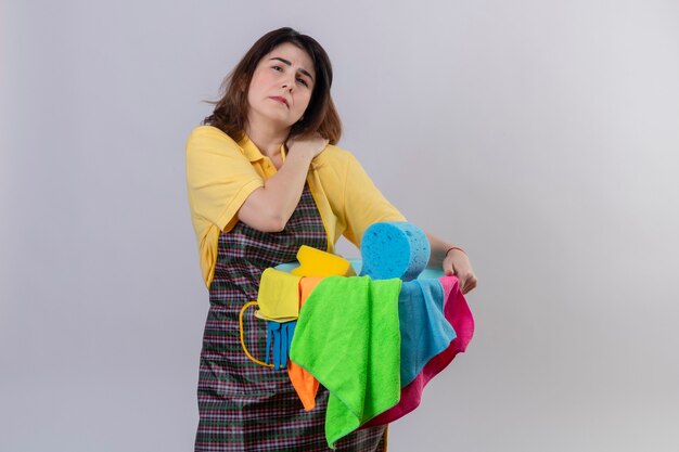 Mujer de mediana edad vistiendo delantal sosteniendo el balde con herramientas de limpieza mirando mal tocando su hombro con dolor de pie sobre la pared blanca