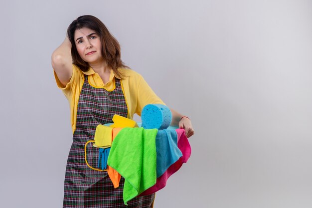 Mujer de mediana edad vistiendo delantal sosteniendo la cuchara con herramientas de limpieza con aspecto cansado y con exceso de trabajo de pie sobre la pared blanca