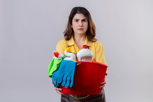 Foto gratuita mujer de mediana edad vistiendo delantal sosteniendo la cuchara con herramientas de limpieza mirando disgustado con el ceño fruncido de pie sobre la pared blanca