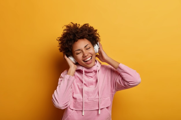 La mujer meloman alegre y relajada escucha música a través de auriculares, cierra los ojos y se siente optimista, vestida con una sudadera con capucha informal, disfruta de un sonido agradable, posa contra la pared amarilla. Gente, ocio, felicidad