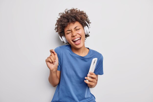 mujer meloman disfruta de su música favorita a través de auriculares sostiene el teléfono móvil tiene movimientos divertidos con el ritmo de la música favorita viste una camiseta azul informal aislada en blanco