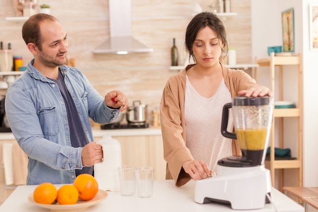 Mujer mezclando frutas en batidora mientras el marido abre la botella de leche. Estilo de vida saludable, despreocupado y alegre, comiendo dieta y preparando el desayuno en una acogedora mañana soleada