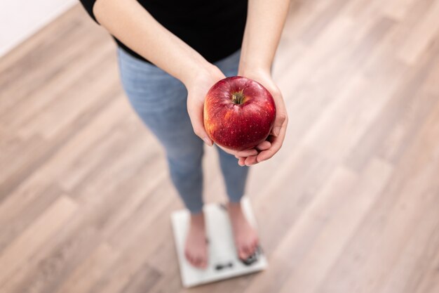 Una mujer mide el peso en una báscula tiene una manzana en sus manos