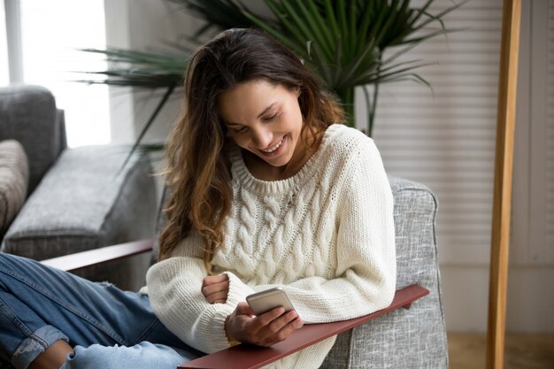 Mujer milenaria feliz sonriente que sostiene el smartphone que se relaja en la butaca