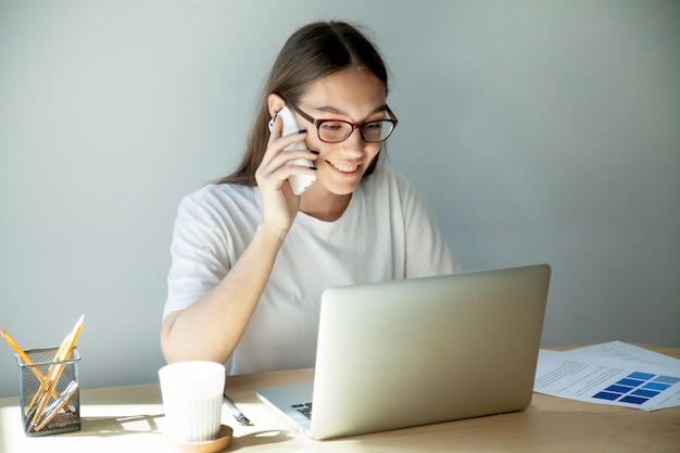 Mujer milenaria en gafas hablando por celular y usando laptop