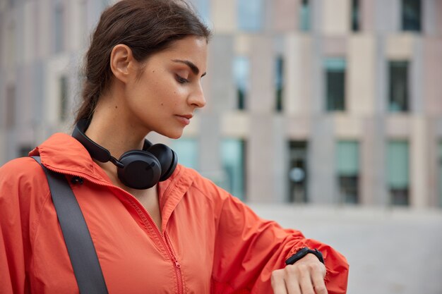 mujer mira actividad rastreador de fitness tiene aplicación de ejecución de pausa de entrenamiento en smartwatch supervisa su actividad deportiva se encuentra al aire libre en la vista borrosa de la ciudad.